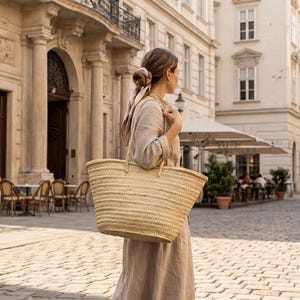 May include: A large, woven straw tote bag with two handles. The bag is a natural beige color and appears to be empty. The bag is being carried by a person wearing a beige dress, walking on a cobblestone street.