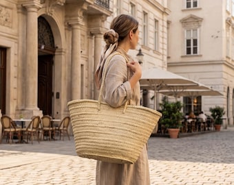May include: A large, woven straw tote bag with two handles. The bag is a natural beige color and appears to be empty. The bag is being carried by a person wearing a beige dress, walking on a cobblestone street.
