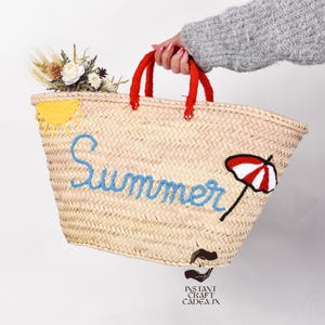 May include: Woven straw tote bag with red handles, embroidered with the word "Summer" in blue, a yellow sun, and a red and white umbrella. The bag is decorated with a small bouquet of flowers.