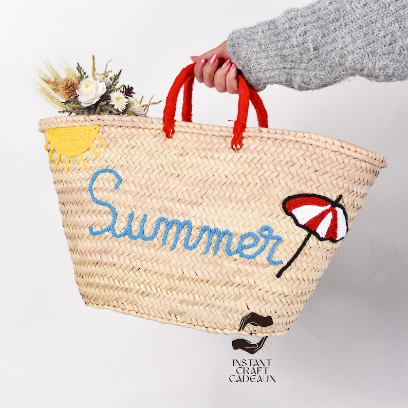 May include: Woven straw tote bag with red handles, embroidered with the word "Summer" in blue, a yellow sun, and a red and white umbrella. The bag is decorated with a small bouquet of flowers.