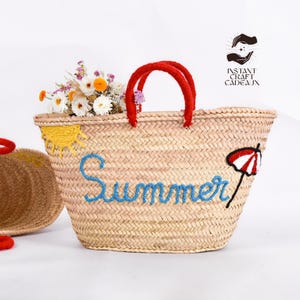 May include: A woven straw tote bag with red handles, embroidered with the word "Summer" in blue. The bag features a yellow sun and a red and white umbrella design. The bag is filled with flowers.