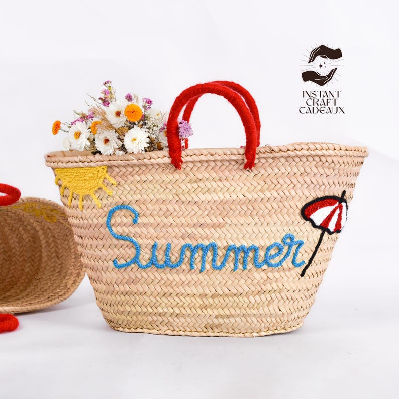 May include: A woven straw tote bag with red handles, embroidered with the word "Summer" in blue. The bag features a yellow sun and a red and white umbrella design. The bag is filled with flowers.