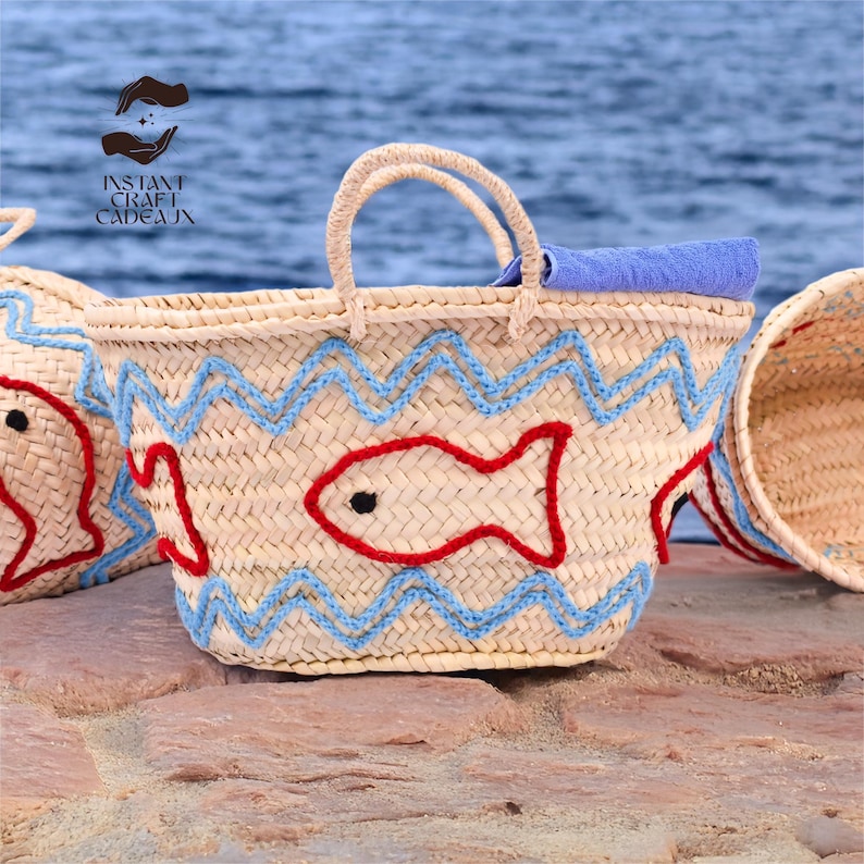May include: Woven straw tote bag with a red fish design and blue zigzag accents. The bag has a light tan color, a pair of handles, and a blue towel inside. The bag is sitting on a stone surface with a blue ocean background.