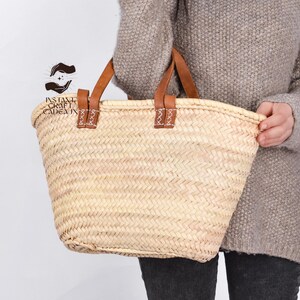 Peut inclure: Un panier en paille tressée avec des anses en cuir marron. Le panier a une forme rectangulaire avec une large ouverture et est tenu par une personne. Les anses sont fixées avec des coutures décoratives. Le panier est de couleur beige naturel.