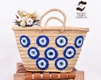 May include: A woven straw tote bag with blue and white crocheted evil eye charms. The bag has a brown leather handle and is sitting on a wooden stool.