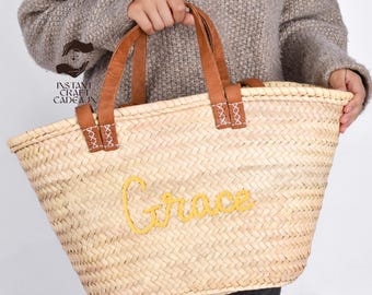 May include: A large, natural woven straw tote bag with brown leather handles. The bag has the name "Grace" embroidered in yellow thread.