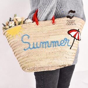 May include: Woven straw tote bag with red handles, embroidered with the word "Summer" in blue. The bag features a yellow sun and a red and white umbrella design. The bag is filled with white and yellow flowers.