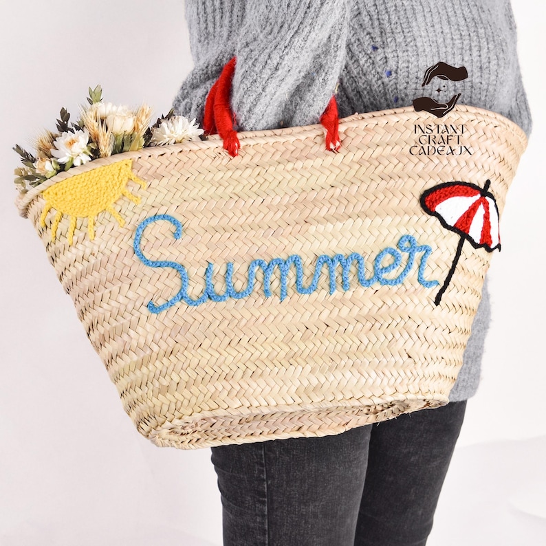 May include: Woven straw tote bag with red handles, embroidered with the word "Summer" in blue. The bag features a yellow sun and a red and white umbrella design. The bag is filled with white and yellow flowers.