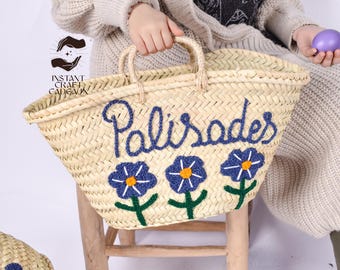 May include: A large, woven straw tote bag with blue embroidered flowers and the word "Palisades" in blue thread. The bag has two handles and is sitting on a wooden chair.