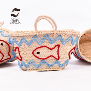 May include: Woven straw tote bags with red fish and blue zigzag embroidery. The bags have beige handles and are set against a white background. The bags are part of the Instant Craft Cadeaux collection.