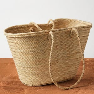 Puede incluir: Un bolso tote de paja tejida con dos asas y un asa de cuerda trenzada. La bolsa es de color marrón claro y se encuentra sobre una superficie marrón rojizo. La bolsa está vacía y tiene una abertura amplia.