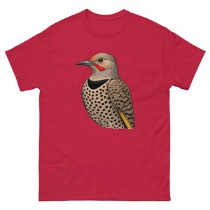 May include: A red short-sleeved t-shirt featuring a centered graphic illustration of a northern flicker bird. The bird has a tan head with a red nape patch, a black crescent on its chest, and tan feathers with black spots and yellow accents.