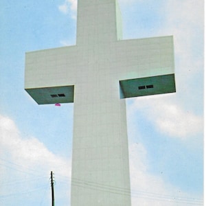 May include: A large, white cross-shaped structure against a blue sky with scattered clouds. The cross has a tall, rectangular base and horizontal arms with openings. The word "PEACE" is visible at the base. A power pole and trees are in the foreground.