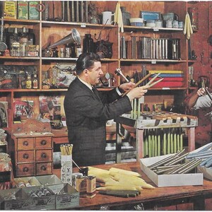May include: Vintage image of a candle shop with a man and woman working. The shop features shelves filled with various items, including jars, boxes, and candles. The man holds candle wicks, while the woman works with a candle mold. A variety of candles are displayed.