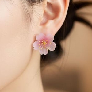 May include: A pair of pink flower-shaped earrings. Each earring has a delicate, translucent petal design with a yellow centre. The earrings are worn on an ear, with the focus on the floral design and its soft, feminine aesthetic.
