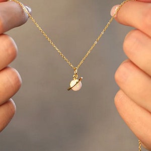 Peut inclure: Un collier en chaîne dorée avec un pendentif délicat en forme de planète en opale. La planète est entourée d'un fin anneau en or.