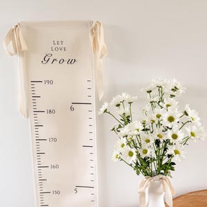 May include: A cream-colored height chart with the words "LET LOVE Grow" printed in black. The chart measures up to 190 cm and is hanging on a white wall. A white vase with white flowers and a wooden surface are also in the image.