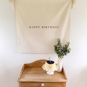 May include: A cream-colored banner with the words "HAPPY BIRTHDAY" in black hangs above a wooden table. A small cake topped with blueberries sits on a white cake stand. A vase of white flowers is to the right.