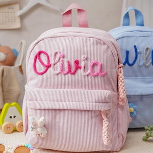 Może przedstawiać: Jasnoróżowy plecak ze sztruksu z wyhaftowanym imieniem Olivia w gradiencie różowej włóczki z przodu. Plecak posiada górny uchwyt, przednią kieszeń zapinaną na zamek z plecionymi uchwytami oraz małą aplikację w kształcie białego króliczka.