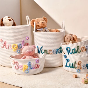 Puede incluir: Un conjunto de cestas de almacenamiento de cuerda tejida blanca con nombres bordados en colores como Angela, Vincent, Sophia, Ralap y Luther. Cada cesta est&aacute; decorada con peque&ntilde;as flores de tela y contiene varios juguetes de peluche.