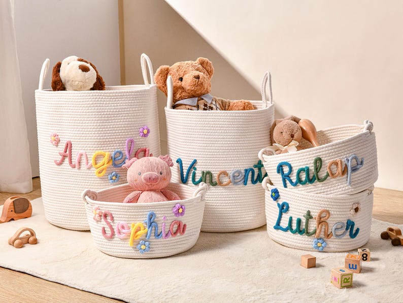 Puede incluir: Un conjunto de cestas de almacenamiento de cuerda tejida blanca con nombres bordados en colores como Angela, Vincent, Sophia, Ralap y Luther. Cada cesta est&aacute; decorada con peque&ntilde;as flores de tela y contiene varios juguetes de peluche.