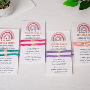 May include: Four cards with rainbow designs and the text "first day of" followed by a grade level. Each card has two bracelets, one larger and one smaller, with a heart charm. The bracelets are in pink, teal, purple, and orange.