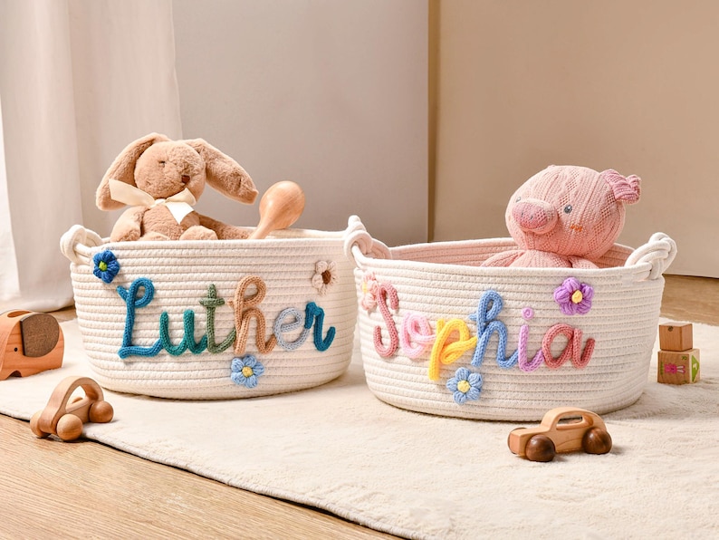 Puede incluir: Dos cestas de almacenamiento redondas de cuerda trenzada color blanco roto con nombres personalizados en letras de lana de colores. Una cesta muestra el nombre Luther en azul y marr&oacute;n, y la otra Sophia en rosa, amarillo y azul, decoradas con flores de lana.