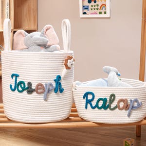 Puede incluir: Dos cestas de almacenamiento de cuerda blanca con asas. La cesta m&aacute;s alta presenta el nombre Joseph en letras de hilo azul, verde y marr&oacute;n con un peque&ntilde;o peluche de le&oacute;n. La cesta m&aacute;s baja presenta el nombre Ralap en colores similares.