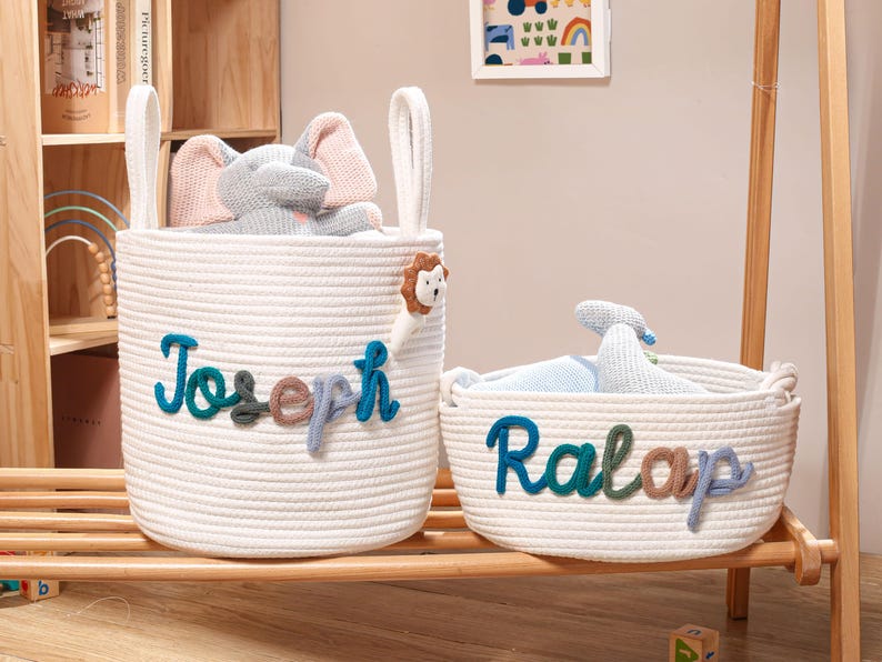 Puede incluir: Dos cestas de almacenamiento de cuerda blanca con asas. La cesta m&aacute;s alta presenta el nombre Joseph en letras de hilo azul, verde y marr&oacute;n con un peque&ntilde;o peluche de le&oacute;n. La cesta m&aacute;s baja presenta el nombre Ralap en colores similares.