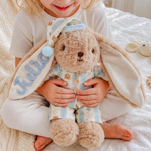 May include: A plush tan rabbit toy wearing light blue and white striped pajamas with small bear patterns and three wooden buttons. The rabbit has a matching nightcap with a light blue pom-pom and the name Julia embroidered in blue script.