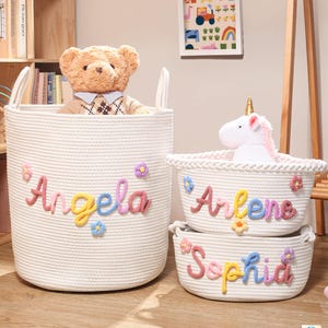 Puede incluir: Tres cestas de almacenamiento de cuerda tejida blanca con asas. Cada cesta presenta un nombre personalizado en letras de hilo coloridas y texturizadas: Angela, Arlene y Sophia. Peque&ntilde;as flores de hilo multicolor est&aacute;n unidas alrededor de cada nombre.