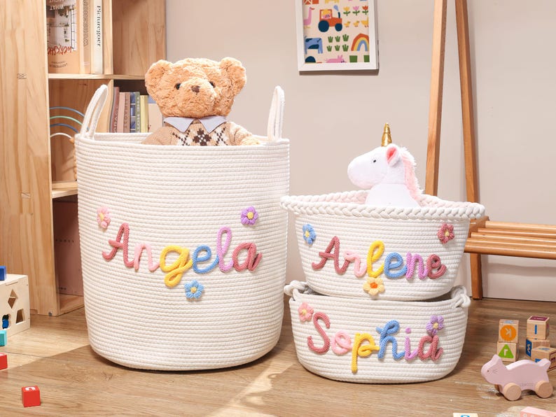 Puede incluir: Tres cestas de almacenamiento de cuerda tejida blanca con asas. Cada cesta presenta un nombre personalizado en letras de hilo coloridas y texturizadas: Angela, Arlene y Sophia. Peque&ntilde;as flores de hilo multicolor est&aacute;n unidas alrededor de cada nombre.