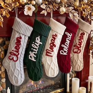 May include: Five cable-knit Christmas stockings hanging in a row. From left to right, the stockings are light gray with red text 'Eileen', dark green with white text 'Philip', cream with red text 'Megan', dark red with white text 'Roland', and beige with red text 'Oliver'.