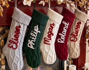 May include: Five cable-knit Christmas stockings hanging in a row. From left to right, the stockings are light grey with red text 'Eileen', dark green with white text 'Philip', cream with red text 'Megan', dark red with white text 'Roland', and beige with red text 'Oliver'.