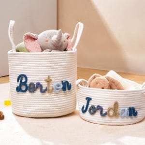Puede incluir: Dos cestas de almacenamiento de cuerda blanca con asas integradas. La cesta m&aacute;s alta presenta el nombre Borton en letras de hilo azul y beige, mientras que la m&aacute;s corta muestra el nombre Jordan en letras de hilo azul y beige, ambas con peluches.
