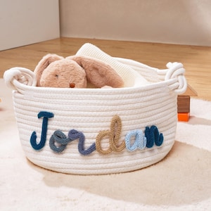 Puede incluir: Una cesta redonda de cuerda de algod&oacute;n color blanco roto con el nombre Jordan en letras de hilo multicolor en la parte frontal. La cesta tiene dos asas laterales y contiene un peluche de color tostado y una manta de punto blanca.