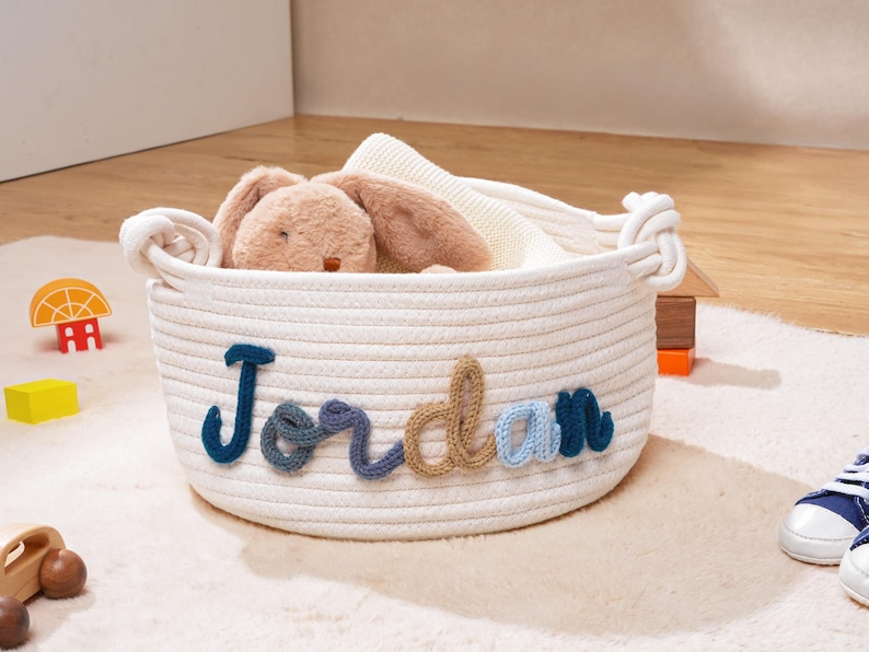 Puede incluir: Una cesta redonda de cuerda de algod&oacute;n color blanco roto con el nombre Jordan en letras de hilo multicolor en la parte frontal. La cesta tiene dos asas laterales y contiene un peluche de color tostado y una manta de punto blanca.