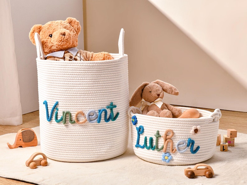 Puede incluir: Dos cestas cil&iacute;ndricas de cuerda blanca con asas integradas. La cesta m&aacute;s alta lleva el nombre Vincent en hilo azul, verde y beige. La cesta m&aacute;s baja lleva el nombre Luther en los mismos colores, decorada con peque&ntilde;as flores azules y blancas.
