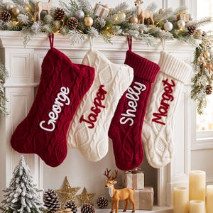 May include: Four cable-knit Christmas stockings hanging in a row, alternating between deep red and cream white. Each stocking features a personalized name embroidered in a contrasting color: George, Jasper, Shelly, and Margot in cursive script.