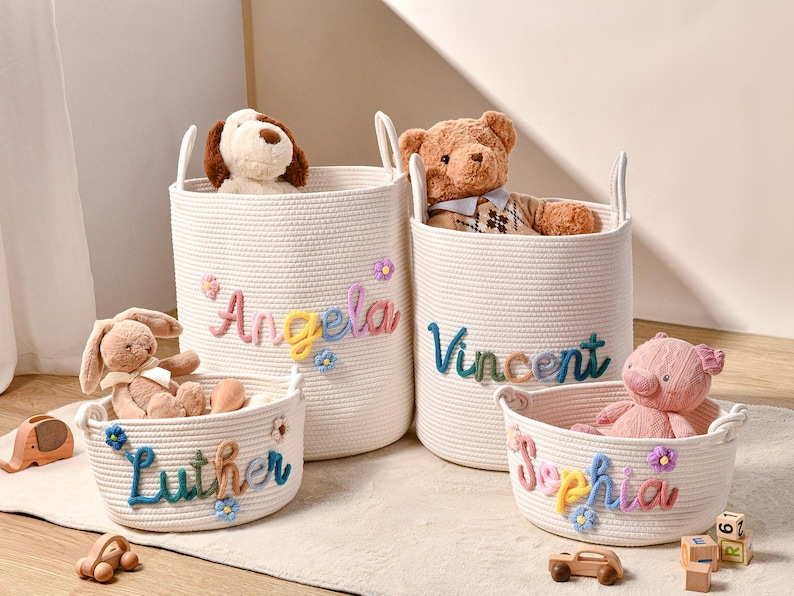 Puede incluir: Cuatro cestas de almacenamiento de cuerda tejida blanca con dos asas cada una. Las cestas presentan nombres bordados de colores: Angela, Vincent, Luther y Sophia, acentuados con peque&ntilde;as flores de tela. Hay peluches dentro de cada cesta.
