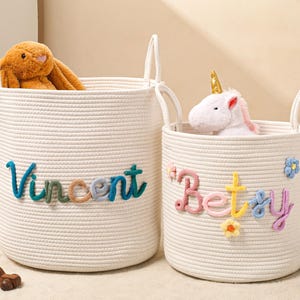 Puede incluir: Dos cestas de almacenamiento cil&iacute;ndricas de cuerda trenzada color blanco roto con asas. La cesta m&aacute;s grande presenta el nombre Vincent en letras de lana multicolor. La cesta m&aacute;s peque&ntilde;a presenta el nombre Betsy en lana con flores.