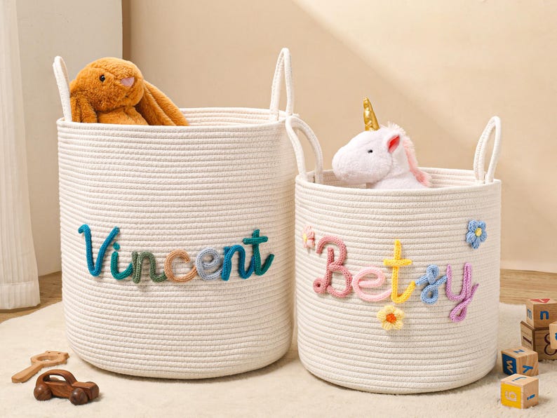 Puede incluir: Dos cestas de almacenamiento cil&iacute;ndricas de cuerda trenzada color blanco roto con asas. La cesta m&aacute;s grande presenta el nombre Vincent en letras de lana multicolor. La cesta m&aacute;s peque&ntilde;a presenta el nombre Betsy en lana con flores.