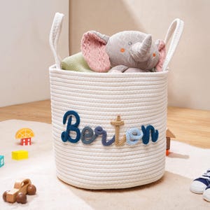 Puede incluir: Una cesta de almacenamiento cil&iacute;ndrica de cuerda blanca con dos asas integradas y el nombre Berton bordado en hilo azul y beige en la parte frontal. La cesta contiene un peluche de elefante gris y un objeto de tela verde, con un dise&ntilde;o limpio.