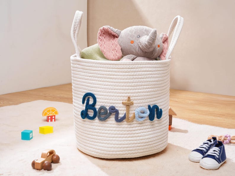 Puede incluir: Una cesta de almacenamiento cil&iacute;ndrica de cuerda blanca con dos asas integradas y el nombre Berton bordado en hilo azul y beige en la parte frontal. La cesta contiene un peluche de elefante gris y un objeto de tela verde, con un dise&ntilde;o limpio.