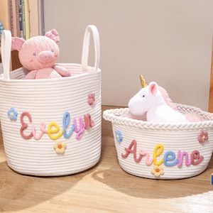 Puede incluir: Dos cestas de almacenamiento de cuerda blanca con asas. La cesta alta lleva el nombre Evelyn en letras de lana de colores con flores, conteniendo un peluche de cerdo rosa. La cesta baja lleva el nombre Arlene con un peluche de unicornio.