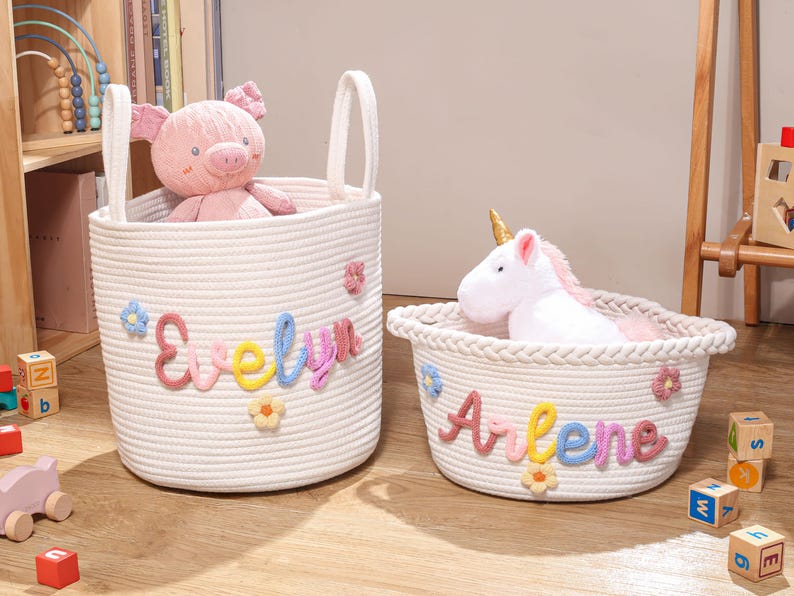 Puede incluir: Dos cestas de almacenamiento de cuerda blanca con asas. La cesta alta lleva el nombre Evelyn en letras de lana de colores con flores, conteniendo un peluche de cerdo rosa. La cesta baja lleva el nombre Arlene con un peluche de unicornio.