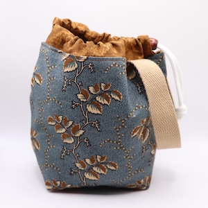 May include: A blue drawstring bag with a floral pattern of brown leaves and vines. The bag has a tan strap and a brown interior. The drawstring is white. The bag is approximately 7 inches tall and 6 inches wide.