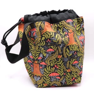 May include: A drawstring bag with a black strap and a black top. The bag features a repeating pattern of red mushrooms, brown tree trunks, green leaves, and snails on a black background. The bag has a wooden bead on the drawstring.