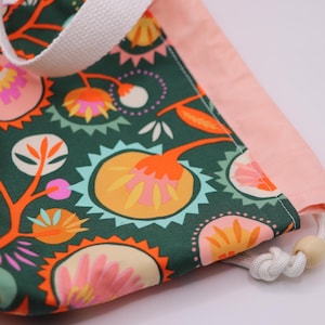 May include: A drawstring bag with a floral pattern in shades of green, orange, pink, and yellow. The bag has a white handle and a light pink border. The drawstring is white with a wooden bead.