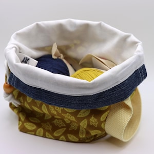 May include: A yellow and blue fabric drawstring bag with a white lining and a denim trim. The bag is filled with two balls of yarn, one blue and one yellow.
